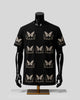A black short-sleeve t-shirt with a repeating beige butterfly and floral pattern, displayed on a black mannequin torso with a gold stand against a light gray background.