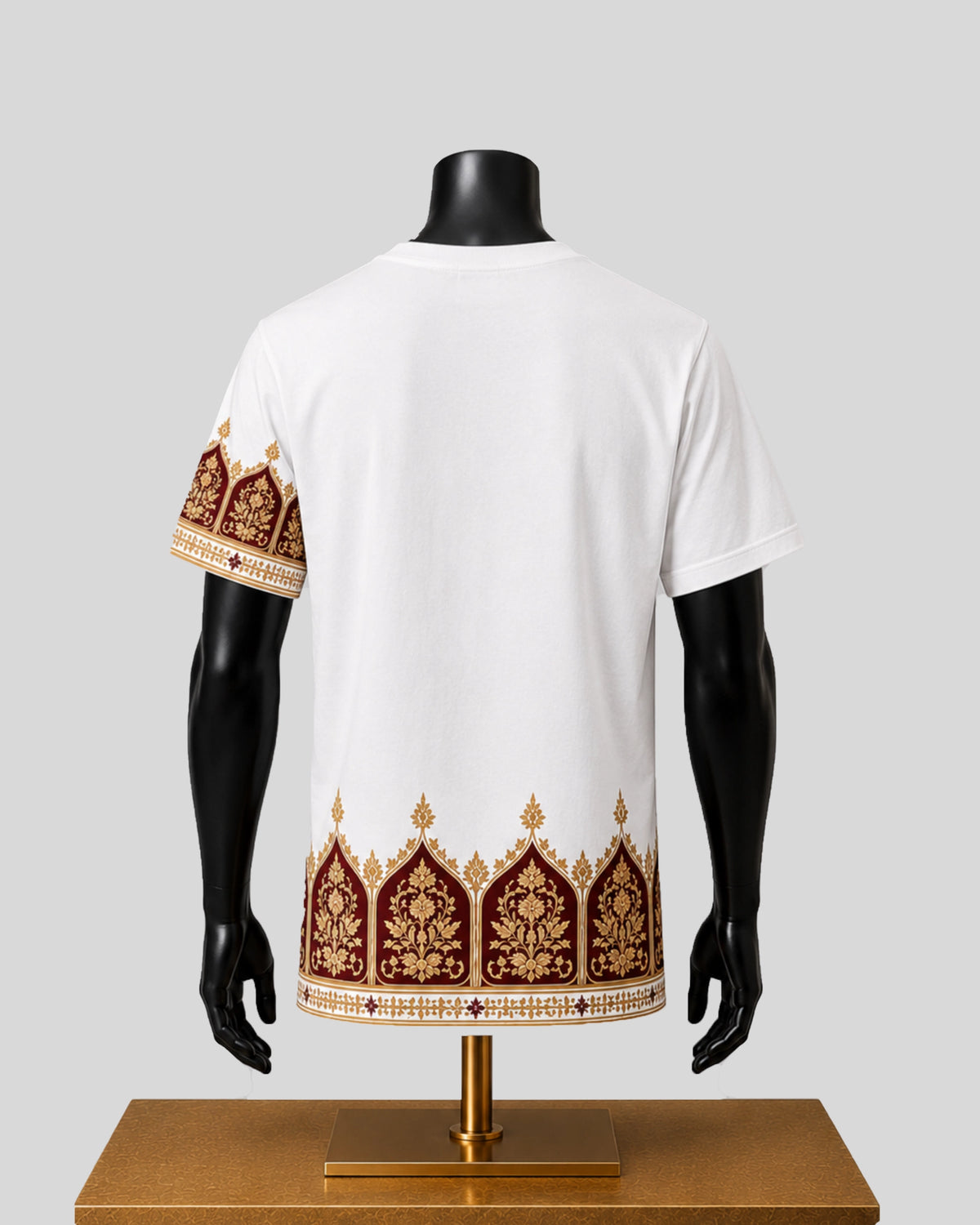 White short-sleeve t-shirt shown from the back on a black mannequin torso, featuring ornate gold and dark red embroidered arch motifs along the bottom hem and left sleeve, displayed on a wooden stand against a light gray background.