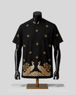 A black short-sleeve t-shirt displayed on a black mannequin torso with a gold stand, featuring gold embroidered floral motifs and a large peacock design across the lower front.