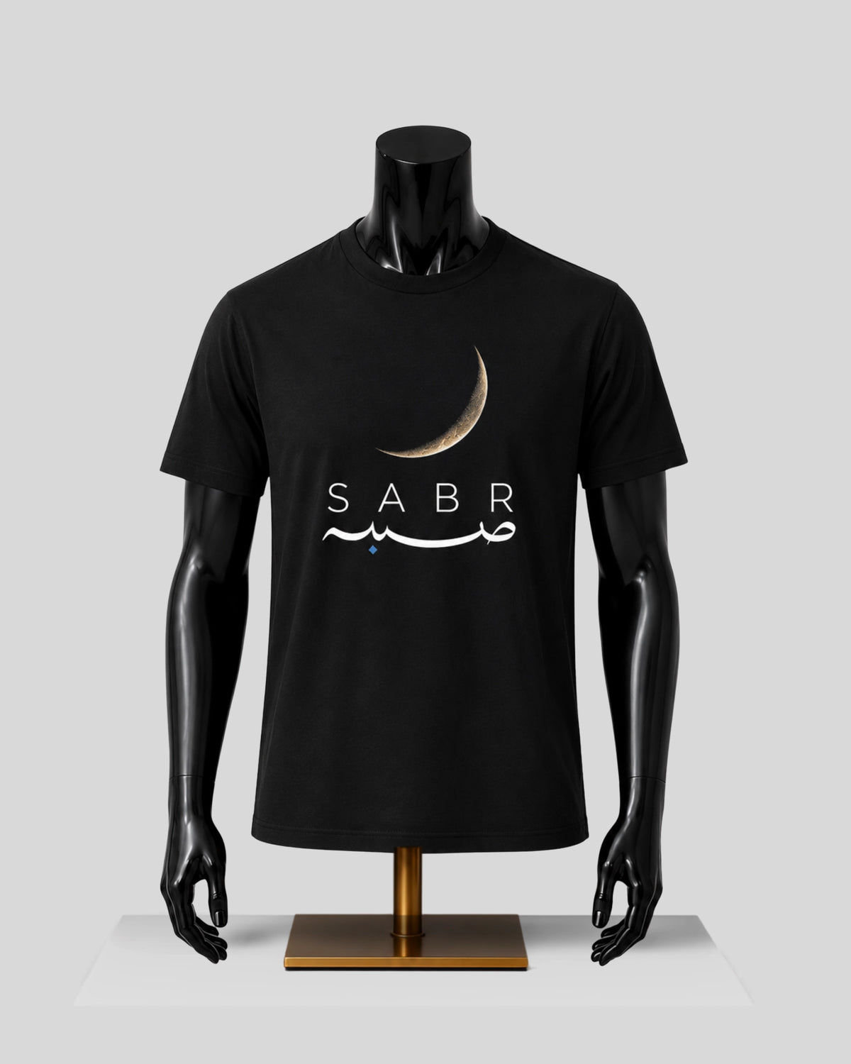 A black short-sleeve t-shirt displayed on a black mannequin torso with arms and hands, standing on a gold-colored base against a light gray background. The shirt features a graphic print of a gold crescent moon above the word 'SABR' in white, with white Arabic calligraphy underneath.