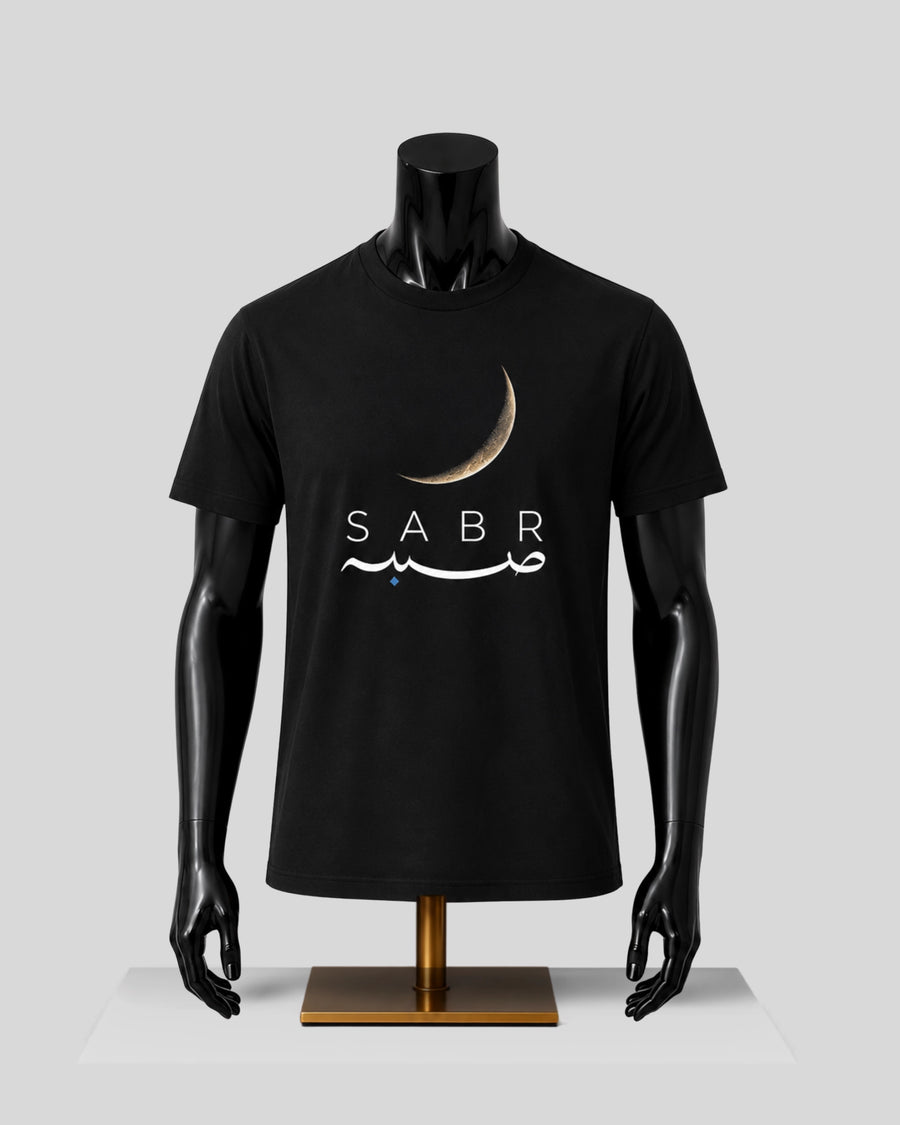 A black short-sleeve t-shirt displayed on a black mannequin torso with arms and hands, standing on a gold-colored base against a light gray background. The shirt features a graphic print of a gold crescent moon above the word 'SABR' in white, with white Arabic calligraphy underneath.