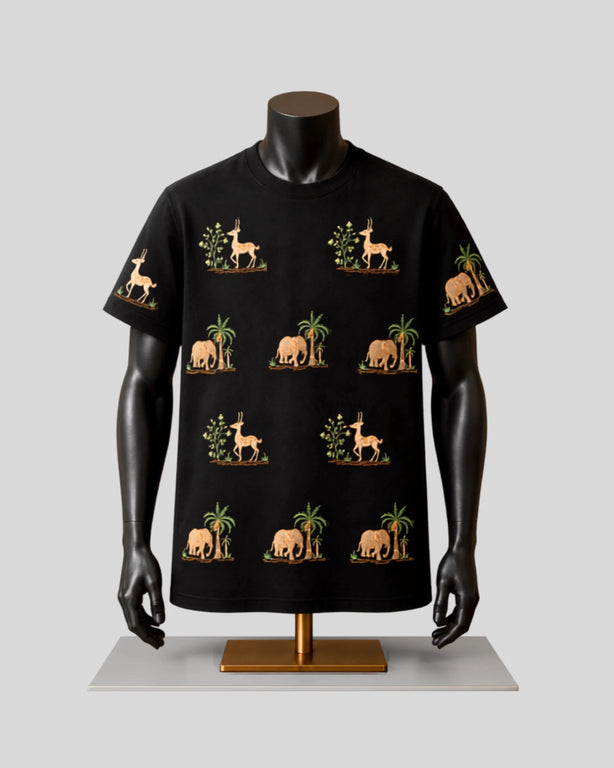 A black short-sleeve t-shirt displayed on a black mannequin torso with arms, featuring a repeating pattern of beige elephants, giraffes, and palm trees with green foliage, set against a light gray background.