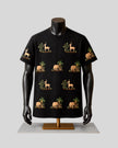 A black short-sleeve t-shirt displayed on a black mannequin torso with arms, featuring a repeating pattern of beige elephants, giraffes, and palm trees with green foliage, set against a light gray background.