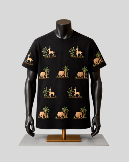 A black short-sleeve t-shirt displayed on a black mannequin torso with arms, featuring a repeating pattern of beige elephants, giraffes, and palm trees with green foliage, set against a light gray background.