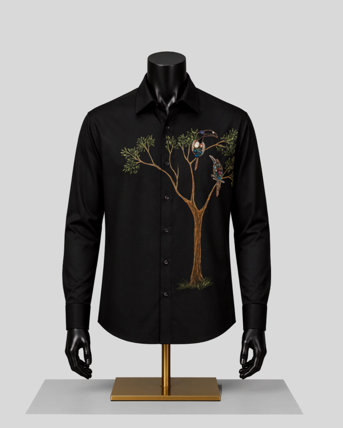 A black long-sleeve button-up shirt displayed on a black mannequin torso with a gold stand, featuring an embroidered tree and two birds on the left chest, against a light gray background.