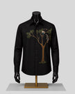 A black long-sleeve button-up shirt displayed on a black mannequin torso with a gold stand, featuring an embroidered tree and two birds on the left chest, against a light gray background.