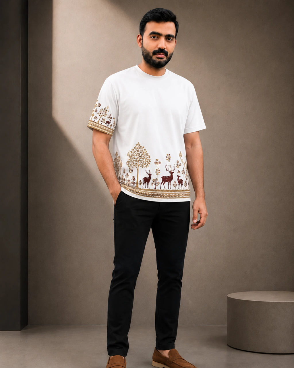 A man wearing a white short-sleeve t-shirt with brown and beige embroidered patterns on the hem and sleeves, paired with black trousers and brown loafers, standing against a neutral gray studio background.