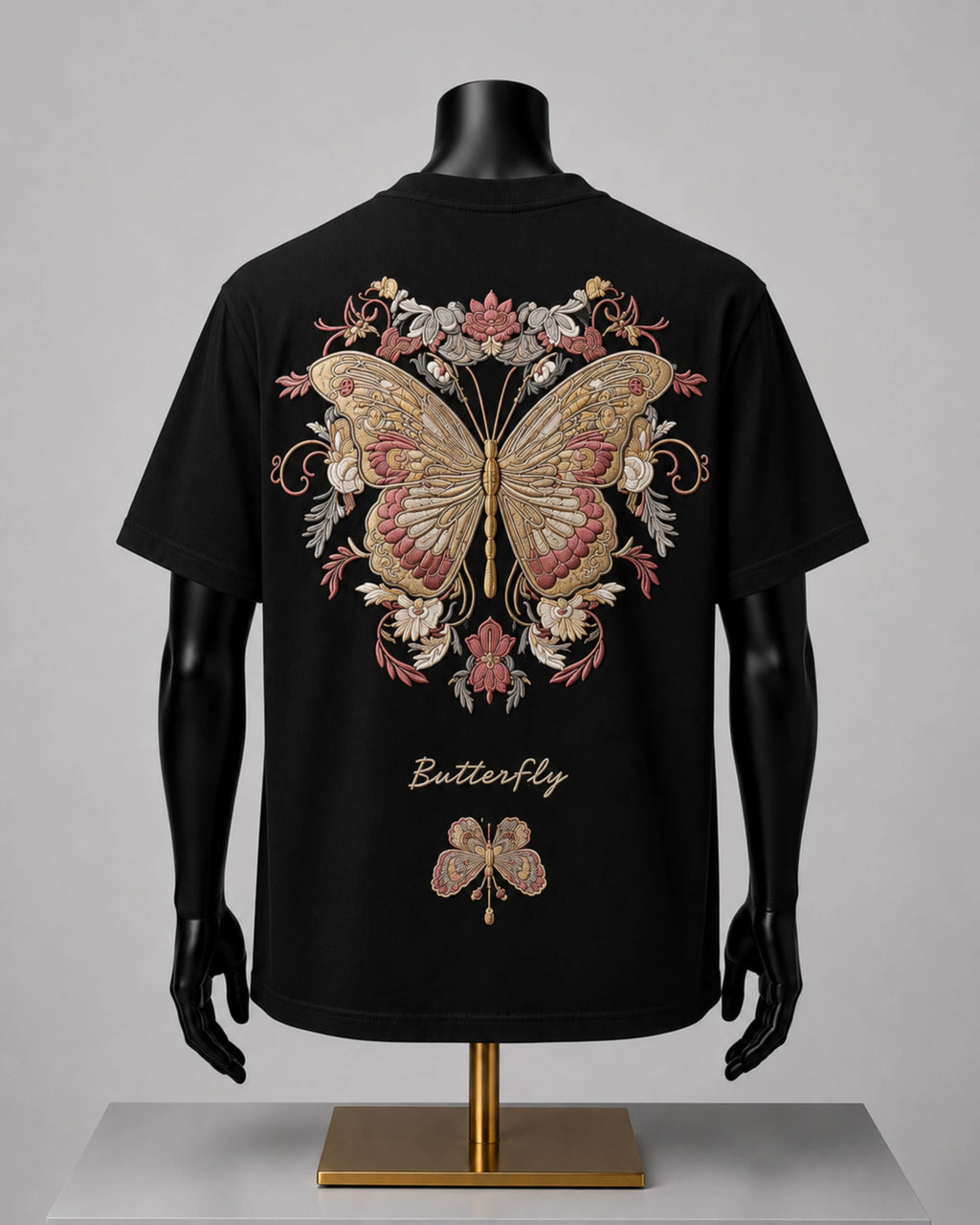 Black short-sleeve T-shirt displayed on a black mannequin torso with arms and hands, shown from the back. The shirt features a large ornate butterfly and floral graphic in beige, pink, and gold tones, with the word 'Butterfly' printed below in a gold script.