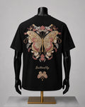 Black short-sleeve T-shirt displayed on a black mannequin torso with arms and hands, shown from the back. The shirt features a large ornate butterfly and floral graphic in beige, pink, and gold tones, with the word 'Butterfly' printed below in a gold script.