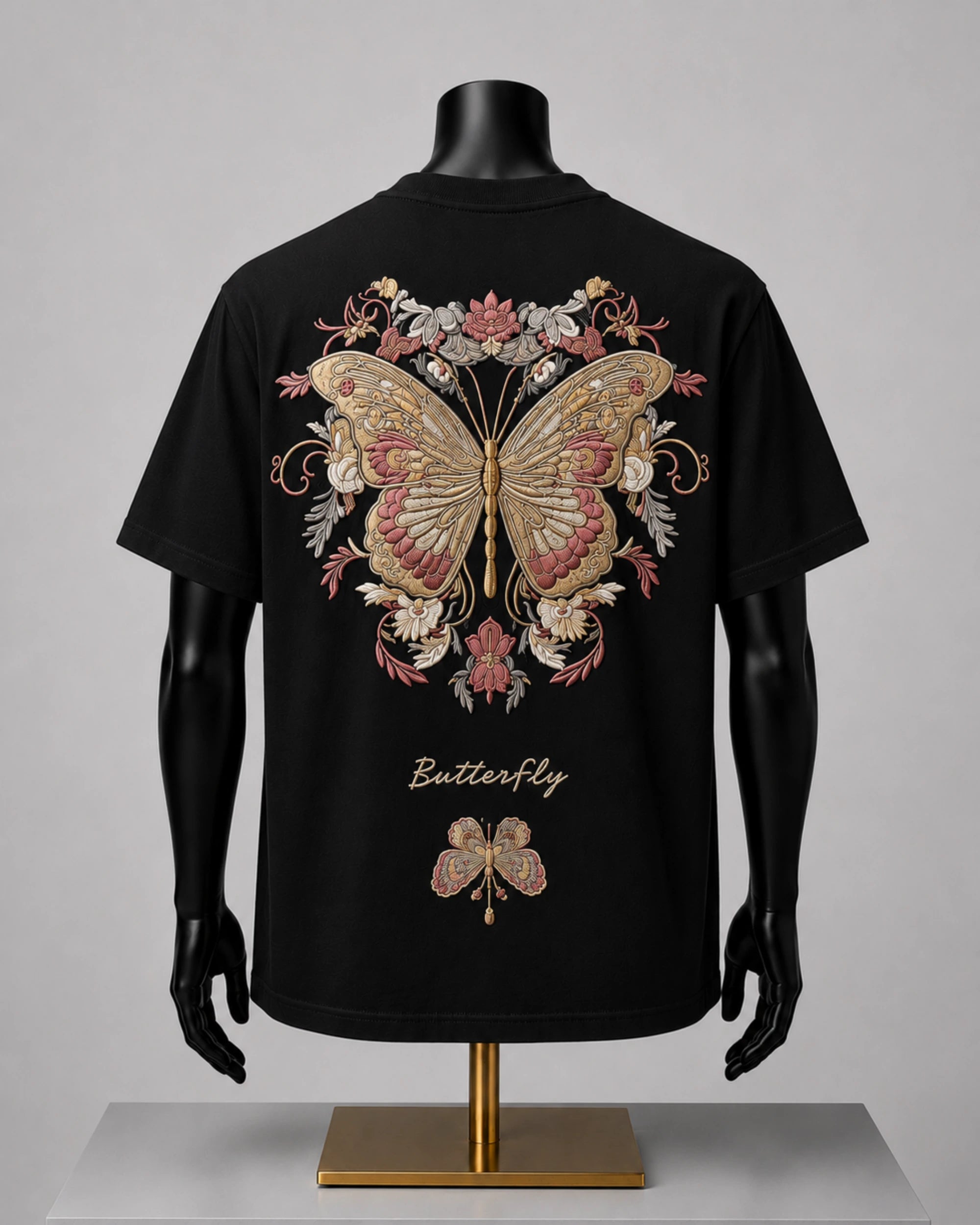 Black short-sleeve T-shirt displayed on a black mannequin torso with arms and hands, shown from the back. The shirt features a large ornate butterfly and floral graphic in beige, pink, and gold tones, with the word 'Butterfly' printed below in a gold script.