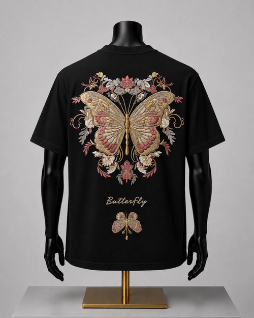 Black short-sleeve T-shirt displayed on a black mannequin torso with arms and hands, shown from the back. The shirt features a large ornate butterfly and floral graphic in beige, pink, and gold tones, with the word 'Butterfly' printed below in a gold script.