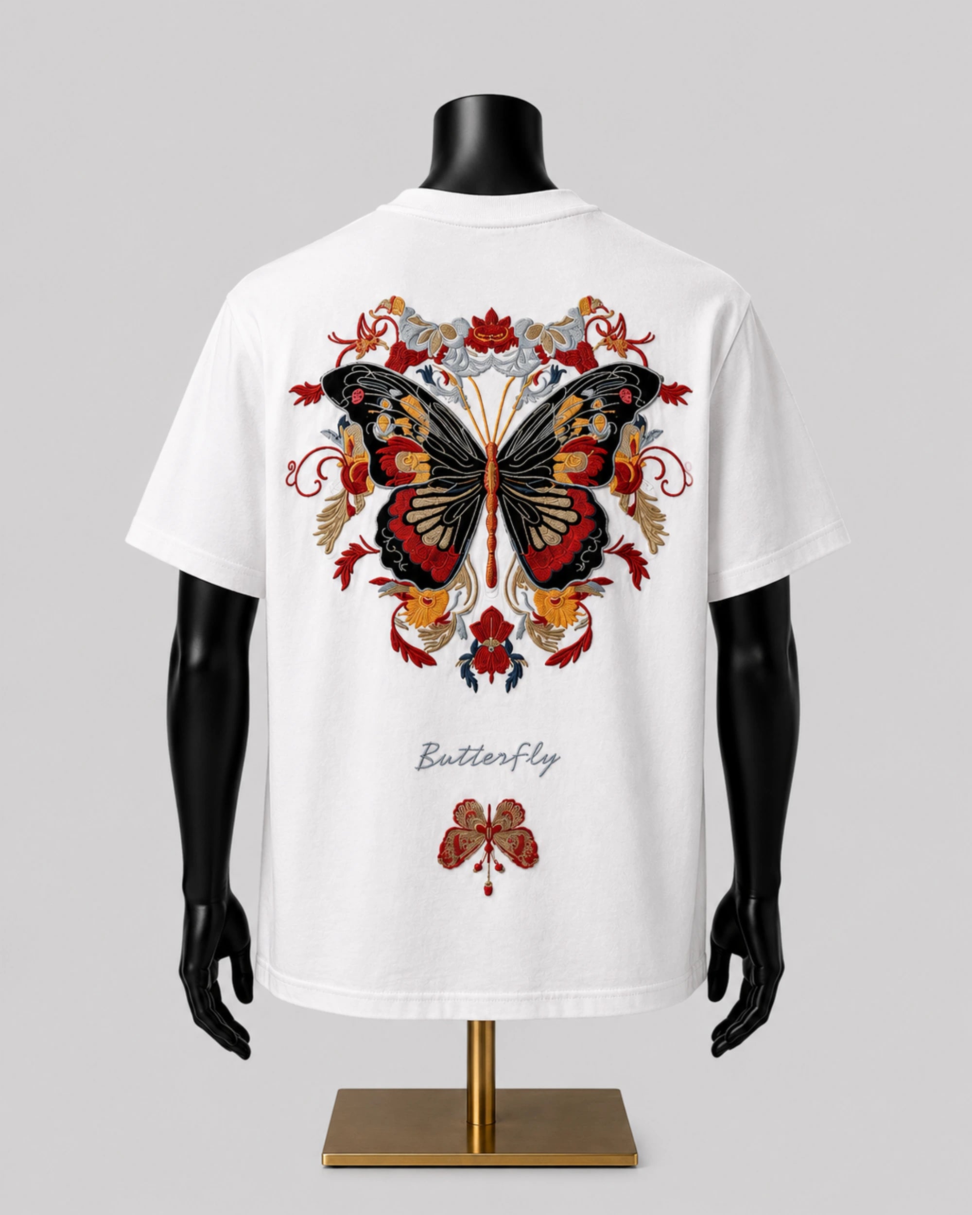 White short-sleeve T-shirt displayed on a black mannequin torso with a gold stand, showing a large butterfly graphic in red, black, and gold tones on the back, with the word 'Butterfly' printed below.