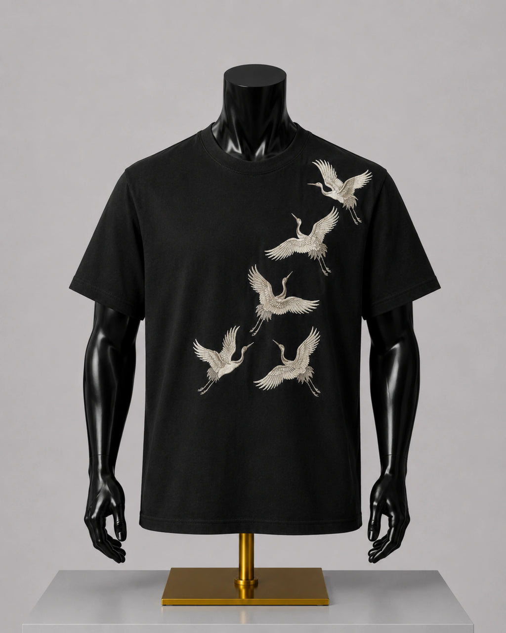 A black short-sleeve crewneck t-shirt displayed on a black mannequin torso with a gold stand, featuring a graphic print of six white cranes flying diagonally across the front.