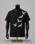 A black short-sleeve crewneck t-shirt displayed on a black mannequin torso with a gold stand, featuring a graphic print of six white cranes flying diagonally across the front.
