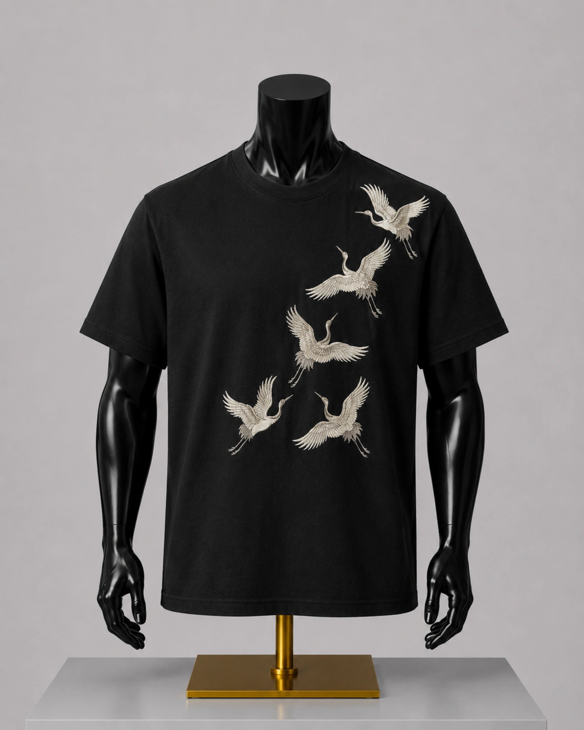 A black short-sleeve crewneck t-shirt displayed on a black mannequin torso with a gold stand, featuring a graphic print of six white cranes flying diagonally across the front.