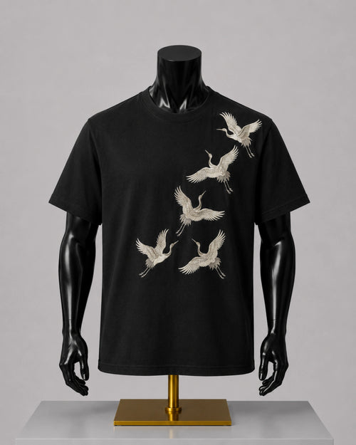 A black short-sleeve crewneck t-shirt displayed on a black mannequin torso with a gold stand, featuring a graphic print of six white cranes flying diagonally across the front.