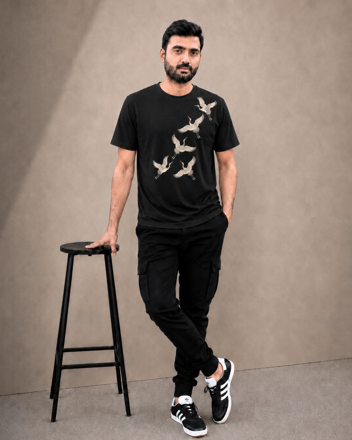 A man wearing a black crew-neck t-shirt with a beige crane graphic print, paired with black cargo pants and black-and-white sneakers, standing next to a black stool against a textured beige studio backdrop.