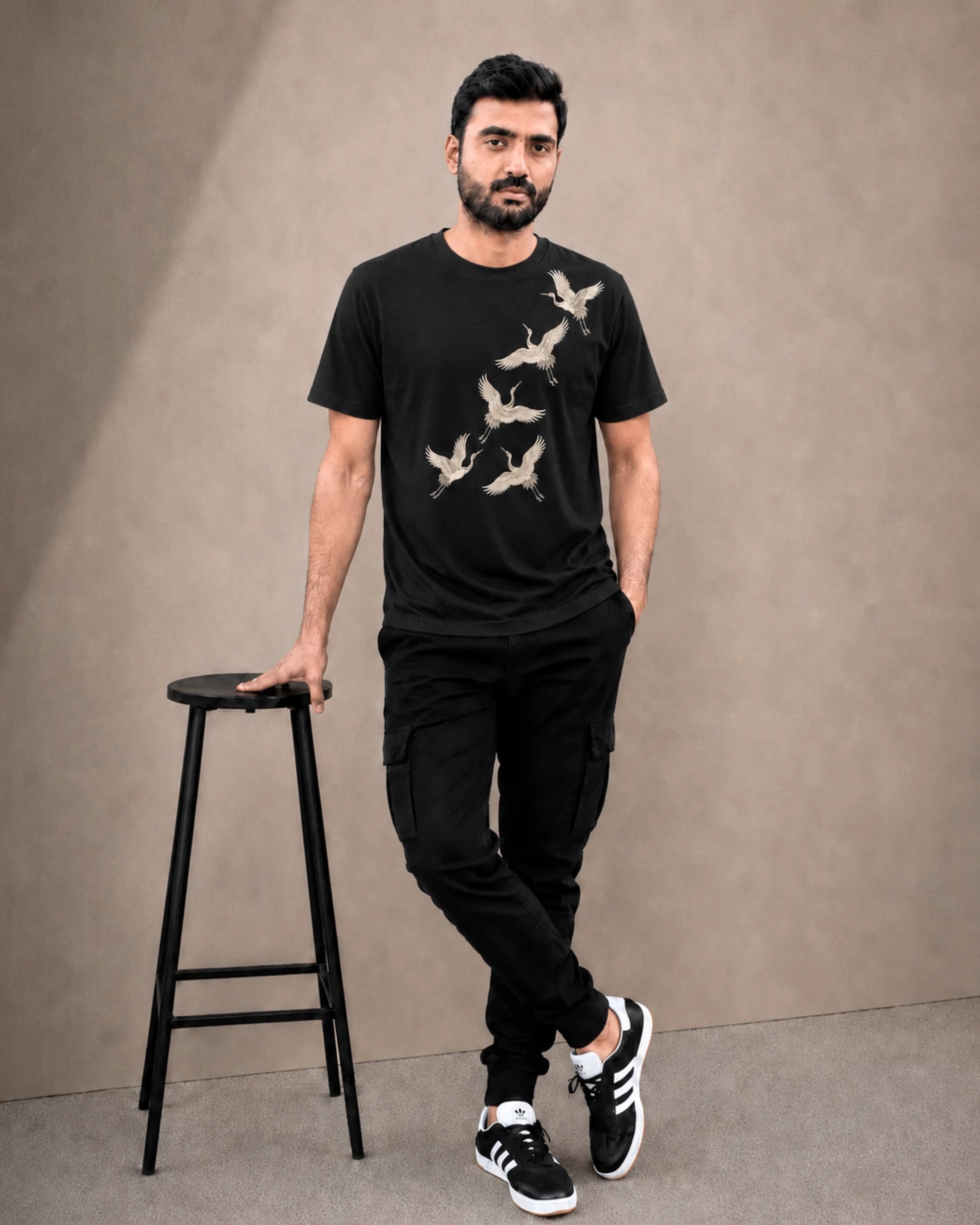 A man wearing a black crew-neck t-shirt with a beige crane graphic print, paired with black cargo pants and black-and-white sneakers, standing next to a black stool against a textured beige studio backdrop.