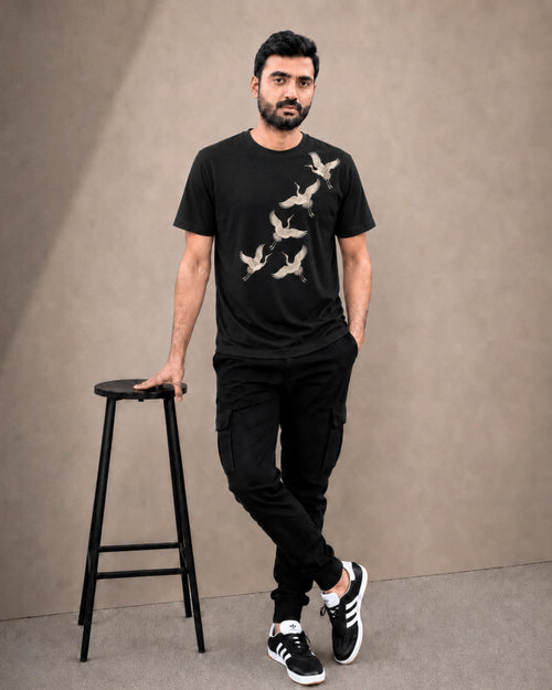 A man wearing a black crew-neck t-shirt with a beige crane graphic print, paired with black cargo pants and black-and-white sneakers, standing next to a black stool against a textured beige studio backdrop.
