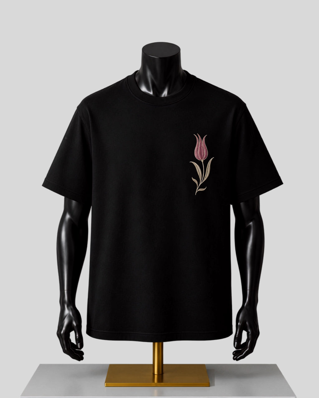 A black short-sleeve crewneck t-shirt displayed on a black mannequin torso with a gold stand, featuring a small pink tulip graphic on the left chest.
