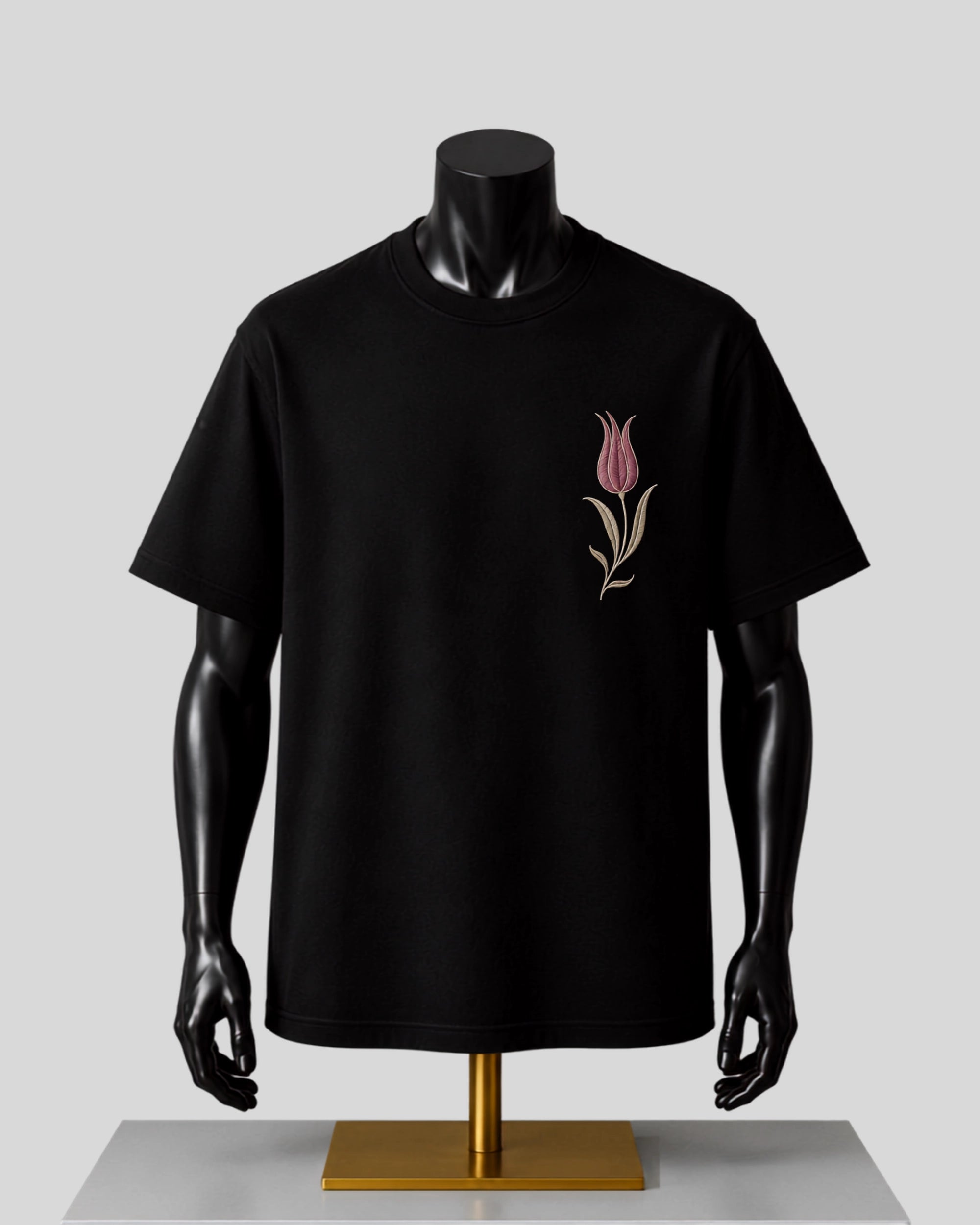 A black short-sleeve crewneck t-shirt displayed on a black mannequin torso with a gold stand, featuring a small pink tulip graphic on the left chest.
