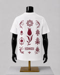 White short-sleeve T-shirt displayed on a black mannequin torso with a gold stand, shown from the back. The shirt features a vertical arrangement of dark red ornamental floral and geometric motifs printed across the back.