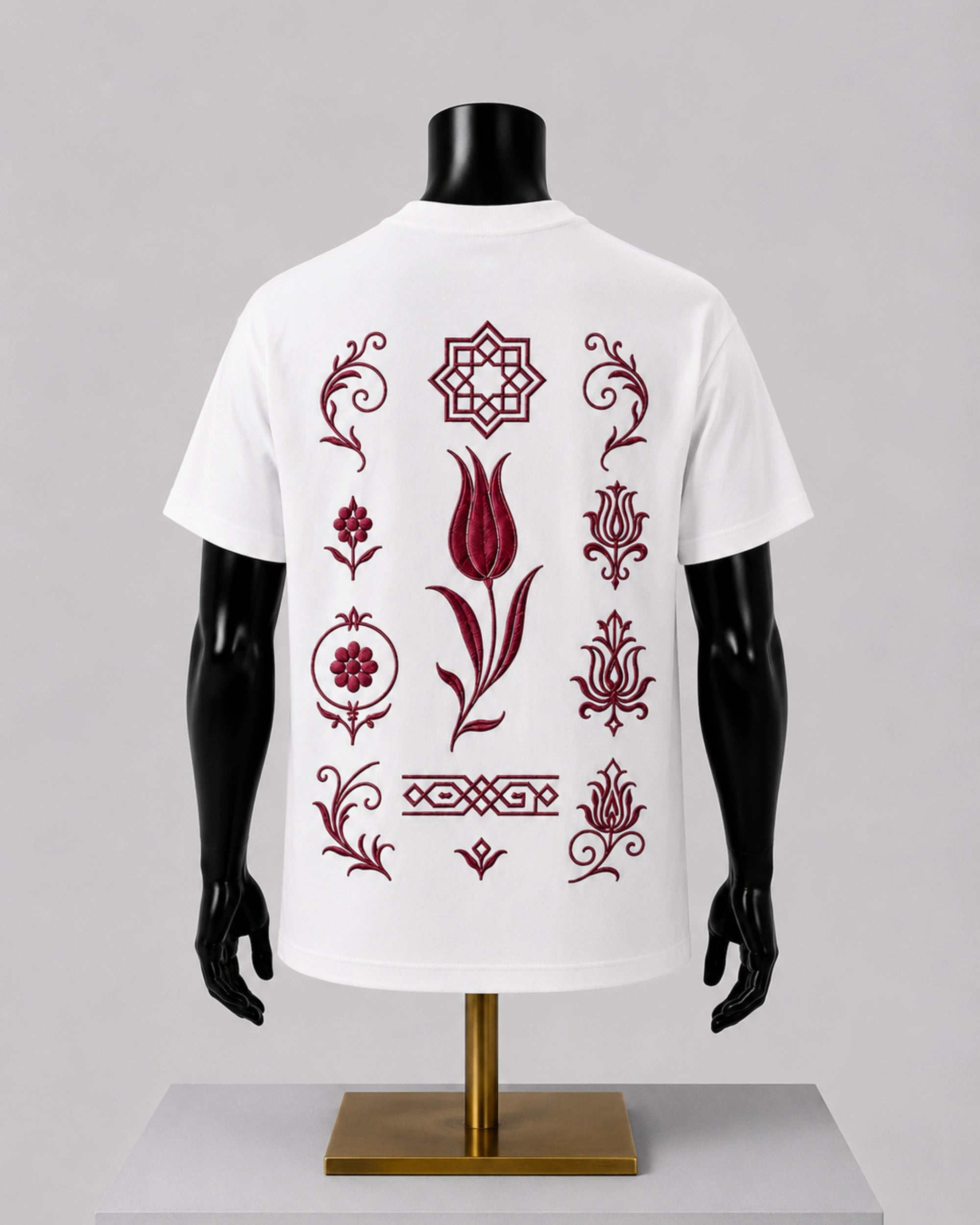 White short-sleeve T-shirt displayed on a black mannequin torso with a gold stand, shown from the back. The shirt features a vertical arrangement of dark red ornamental floral and geometric motifs printed across the back.
