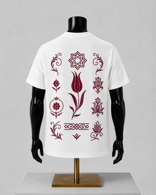 White short-sleeve T-shirt displayed on a black mannequin torso with a gold stand, shown from the back. The shirt features a vertical arrangement of dark red ornamental floral and geometric motifs printed across the back.