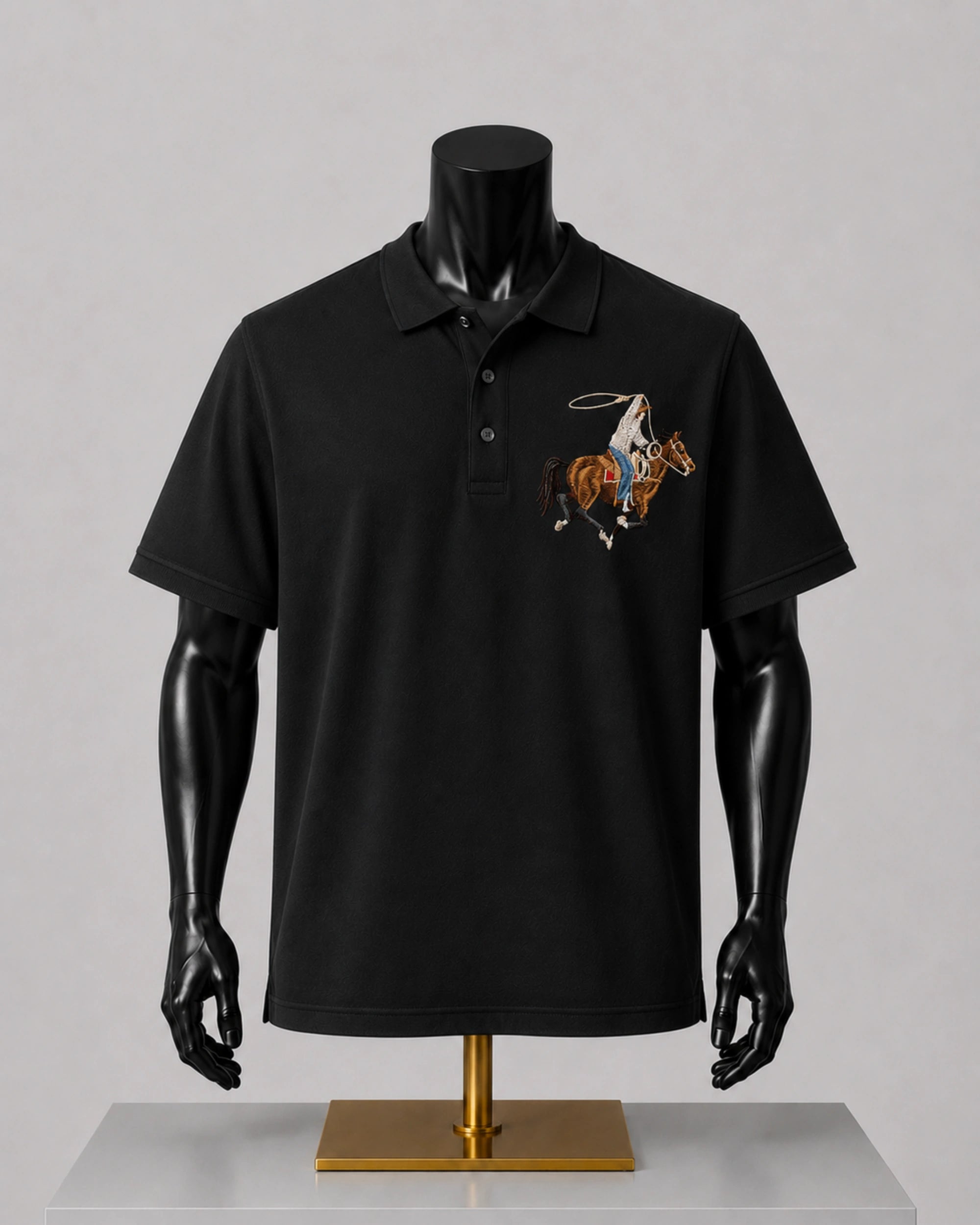 A black short-sleeve polo shirt displayed on a black mannequin torso with a gold stand, featuring a small embroidered cowboy motif on the left chest, set against a neutral gray background.