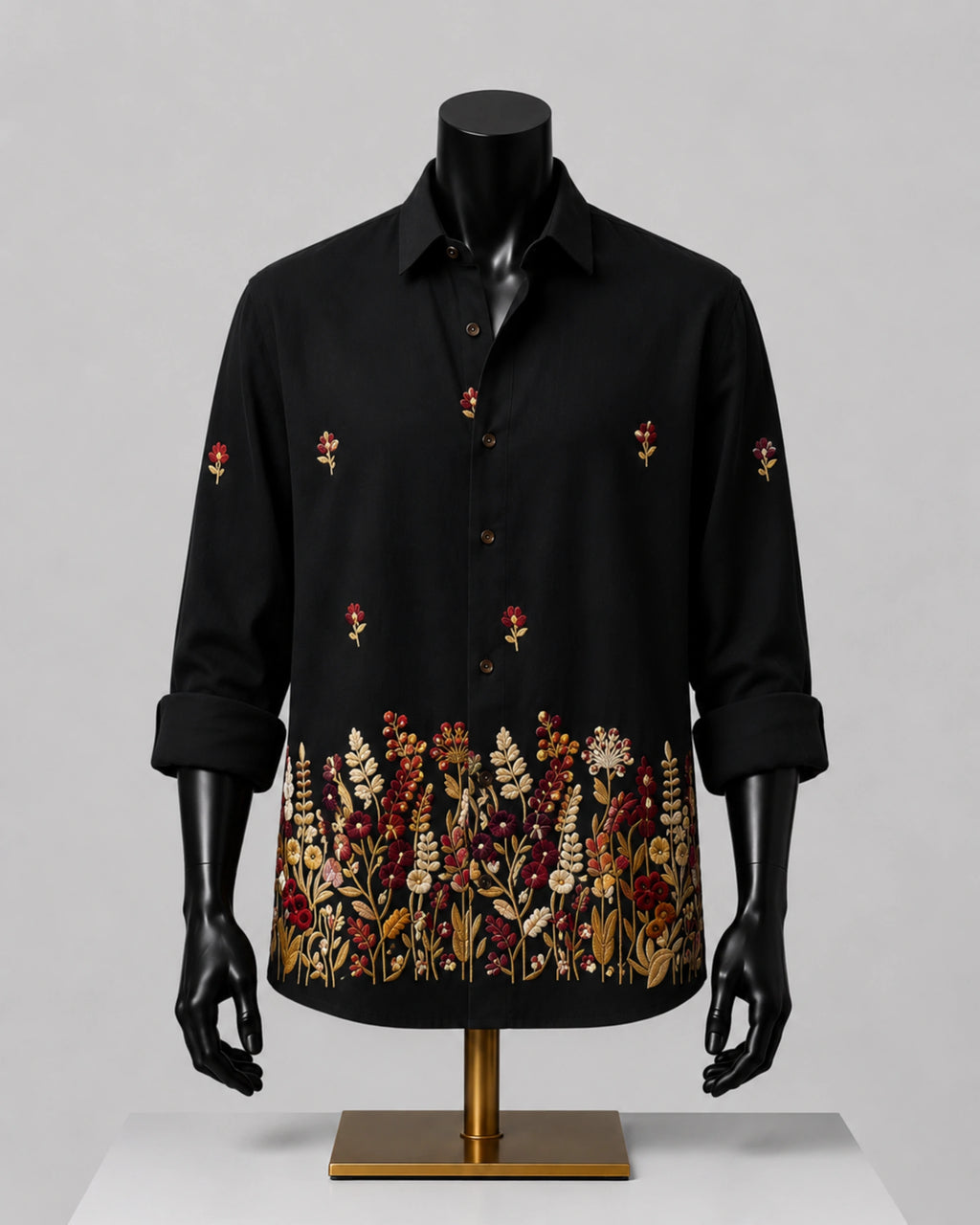 A black long-sleeve button-up shirt displayed on a black mannequin torso with a gold stand, featuring embroidered floral patterns on the front and cuffs, set against a neutral gray background.
