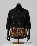 A black long-sleeve button-up shirt displayed on a black mannequin torso with a gold stand, featuring embroidered floral patterns on the front and cuffs, set against a neutral gray background.