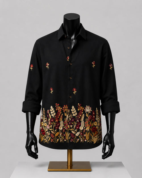 A black long-sleeve button-up shirt displayed on a black mannequin torso with a gold stand, featuring embroidered floral patterns on the front and cuffs, set against a neutral gray background.
