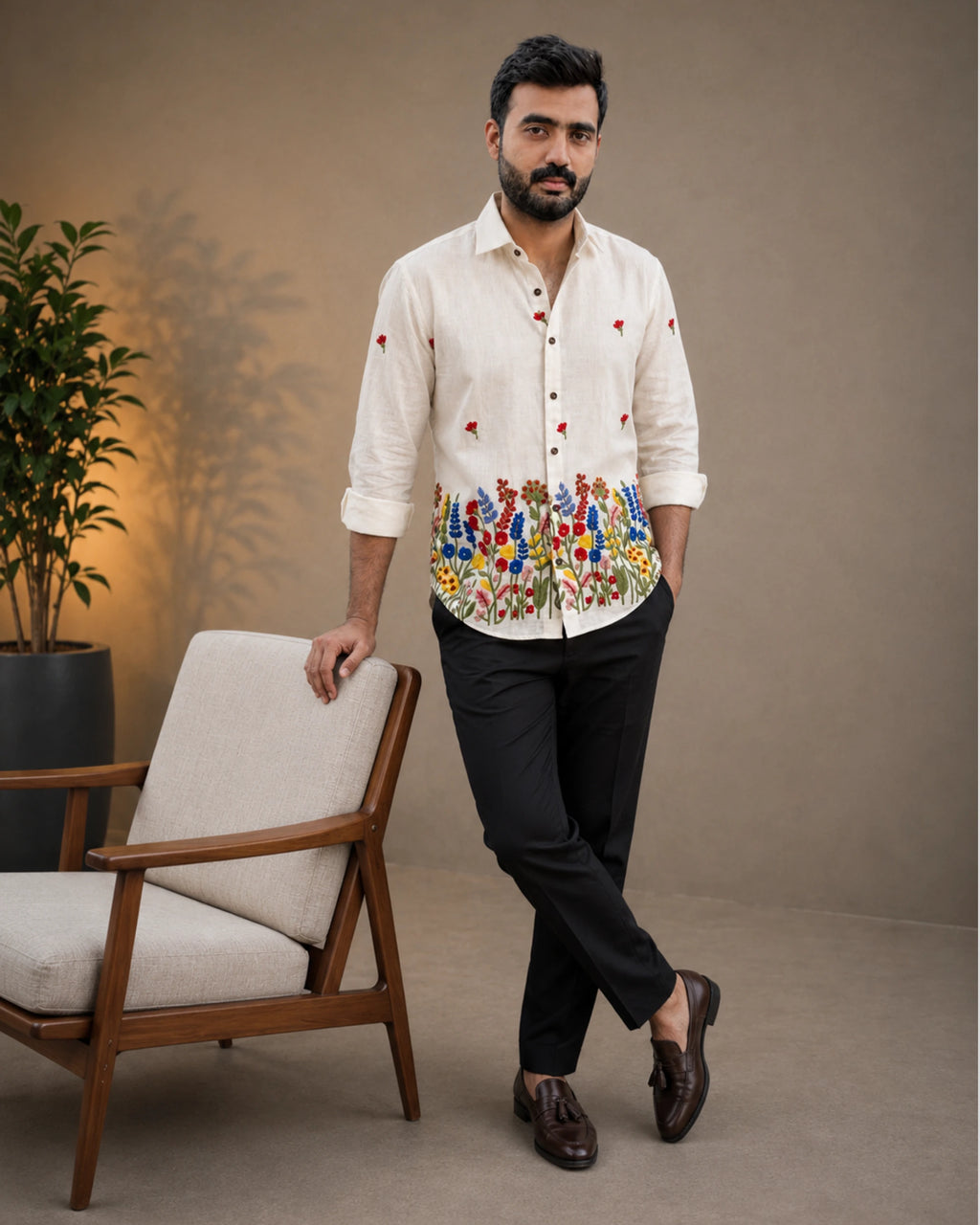 A man wearing a cream-colored embroidered shirt with colorful floral patterns and black trousers, standing next to a modern wooden armchair in a studio setting.