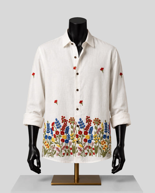 An off-white linen long-sleeve button-up shirt with colorful floral embroidery along the hem and scattered red flower motifs on the sleeves, displayed on a black mannequin torso with a gold stand against a light gray background.