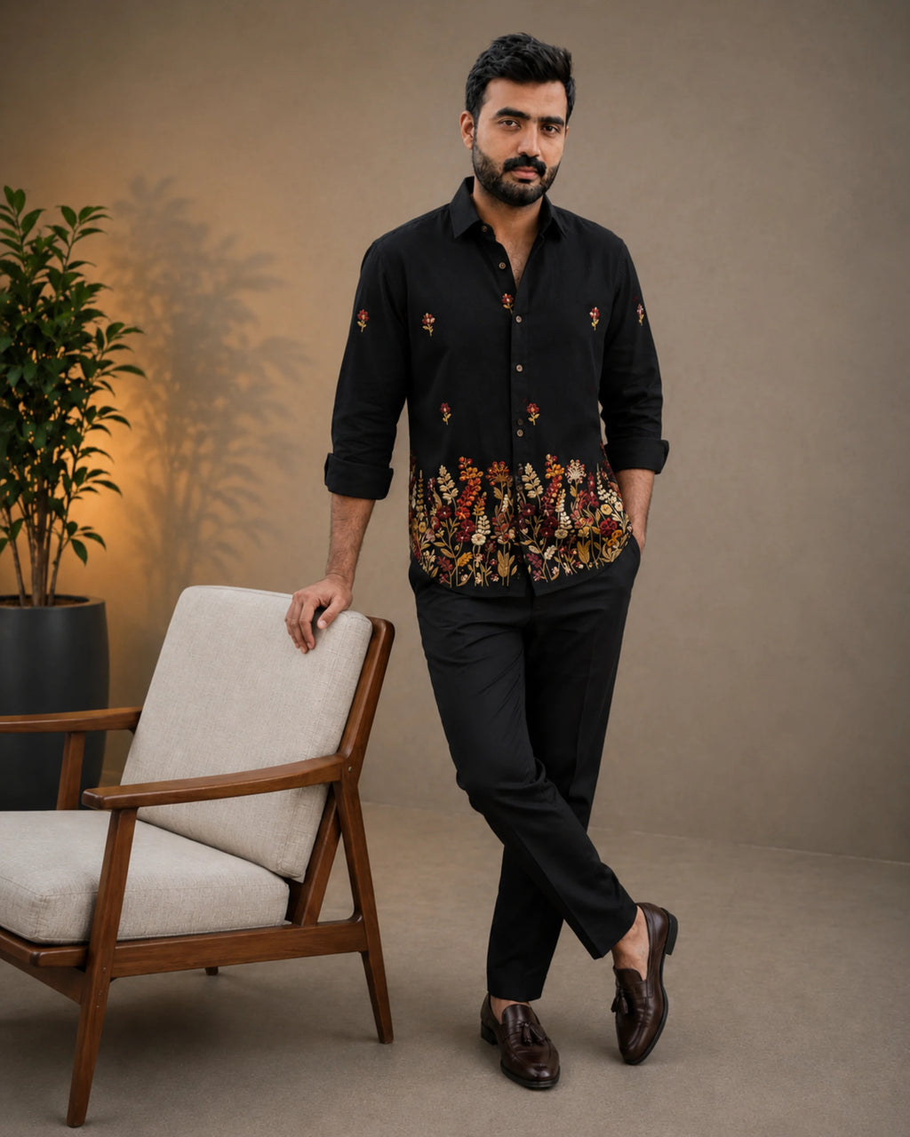 A man wearing a black long-sleeve button-up shirt with colorful floral embroidery concentrated along the hem and scattered on the sleeves, paired with black trousers and dark brown loafers, standing next to a modern armchair in a studio setting.