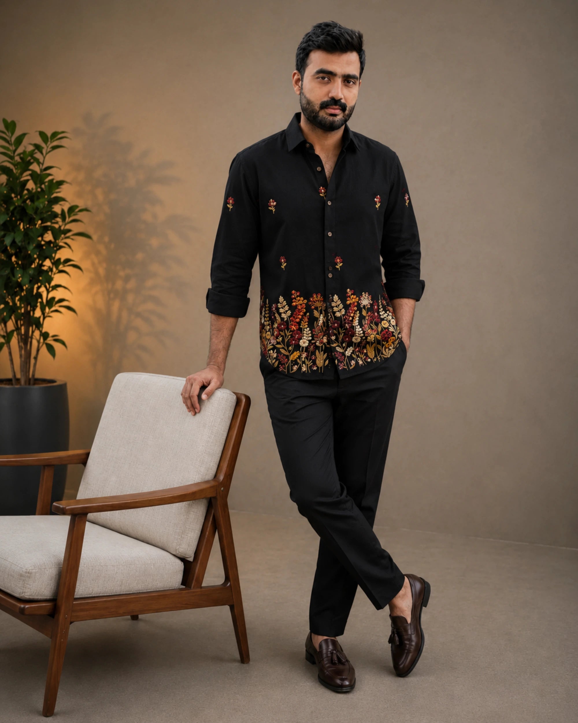 A man wearing a black long-sleeve button-up shirt with colorful floral embroidery concentrated along the hem and scattered on the sleeves, paired with black trousers and dark brown loafers, standing next to a modern armchair in a studio setting.