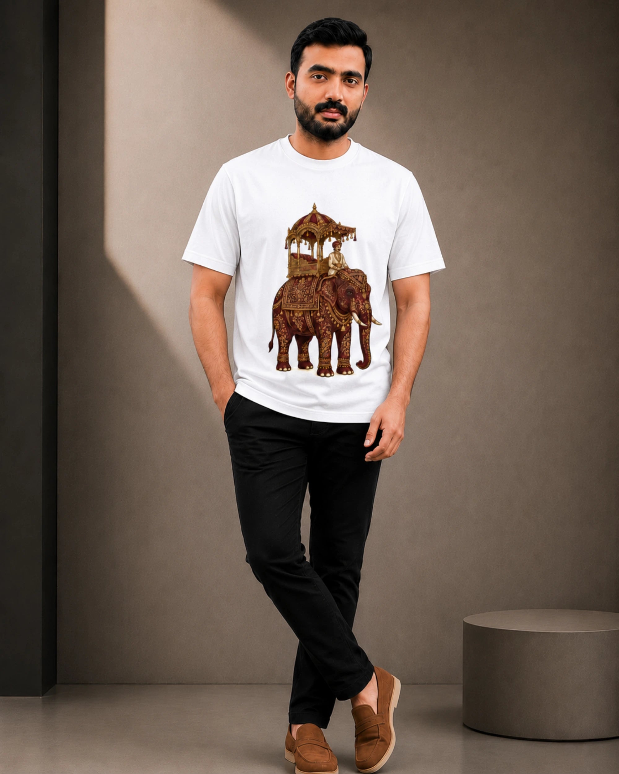 A man wearing a white short-sleeve t-shirt with a brown elephant and palanquin graphic, paired with black pants and brown loafers, standing in a studio setting.