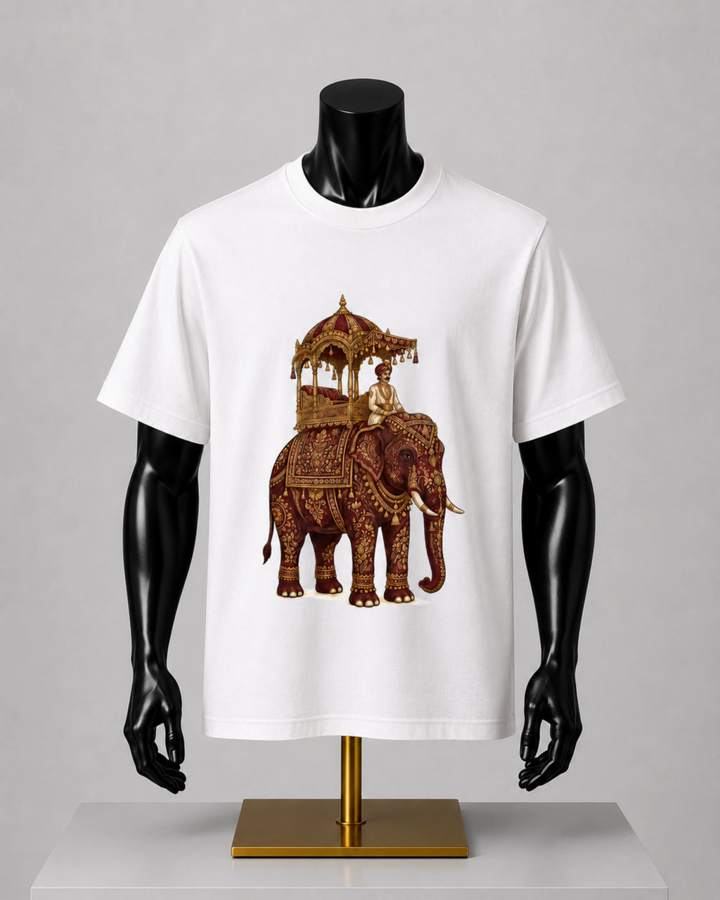 A white short-sleeve crewneck t-shirt displayed on a black mannequin torso. The shirt features a detailed graphic print of an ornate brown and gold decorated elephant carrying a traditional canopy with a rider.