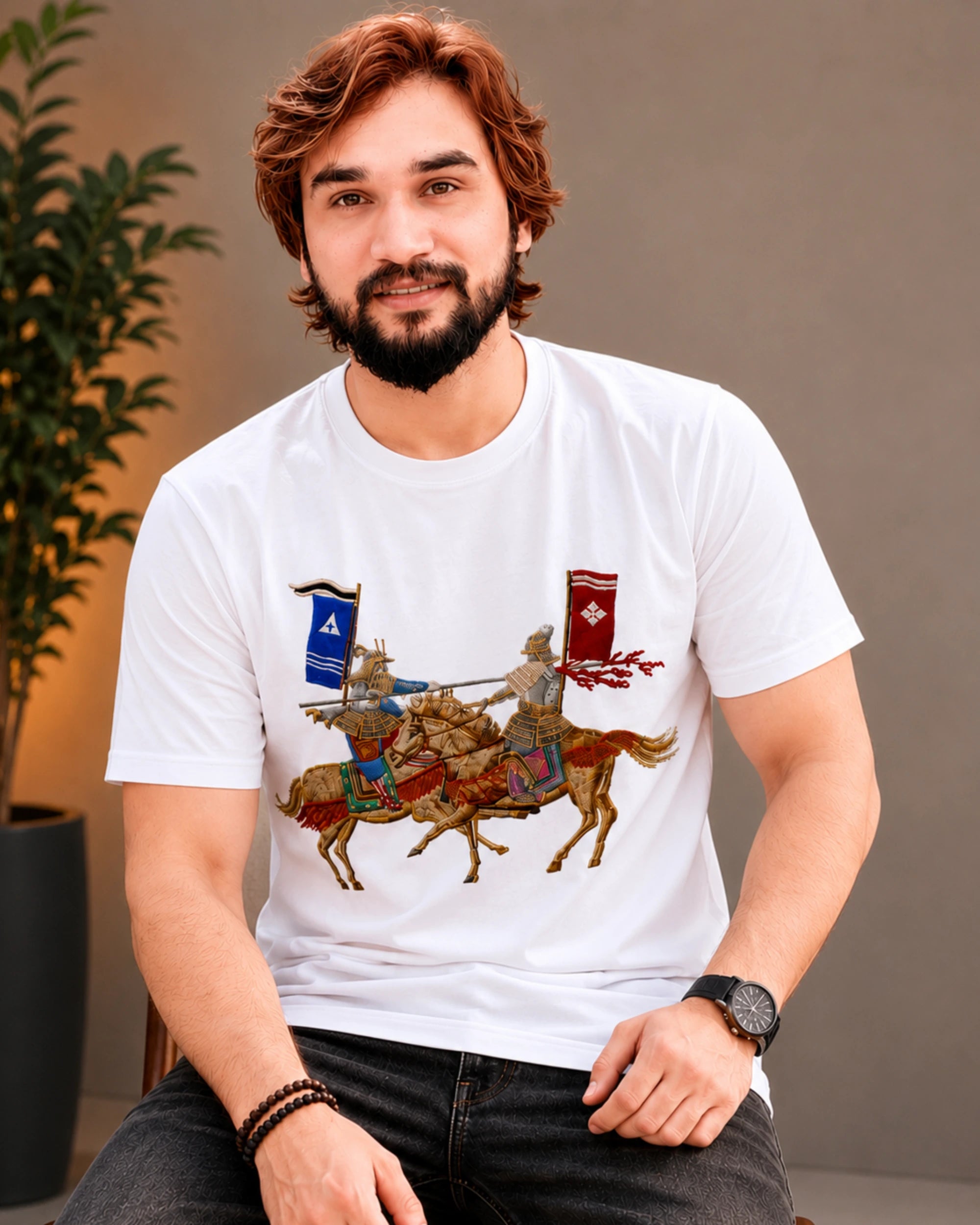 A man wearing a white short-sleeve t-shirt with a detailed graphic of knights on horseback in the center, paired with dark gray jeans, sitting in a styled indoor setting.