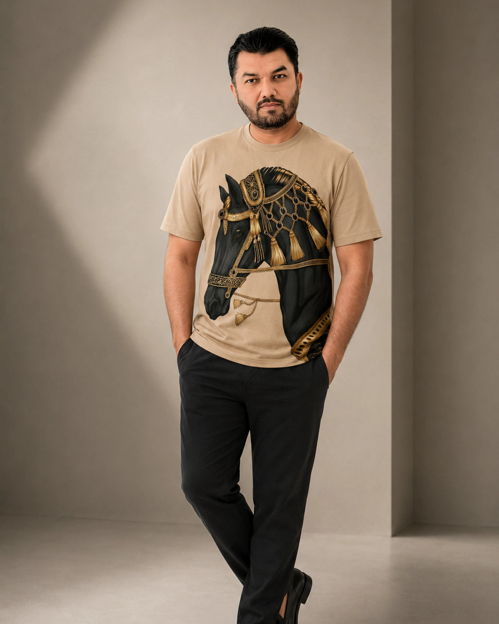 A man wearing a beige t-shirt with a large black and gold horse graphic print, paired with black trousers, standing in a studio with a warm neutral background.