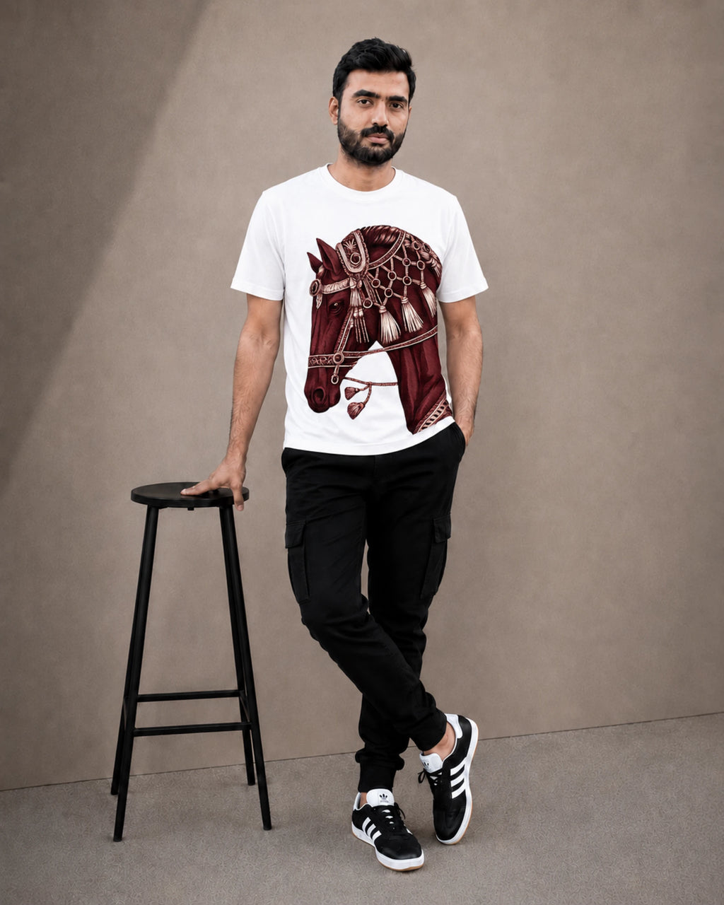 A man wearing a white crew-neck t-shirt with a detailed maroon horse graphic print, paired with black cargo pants and black-and-white sneakers, standing next to a black stool against a neutral studio backdrop.