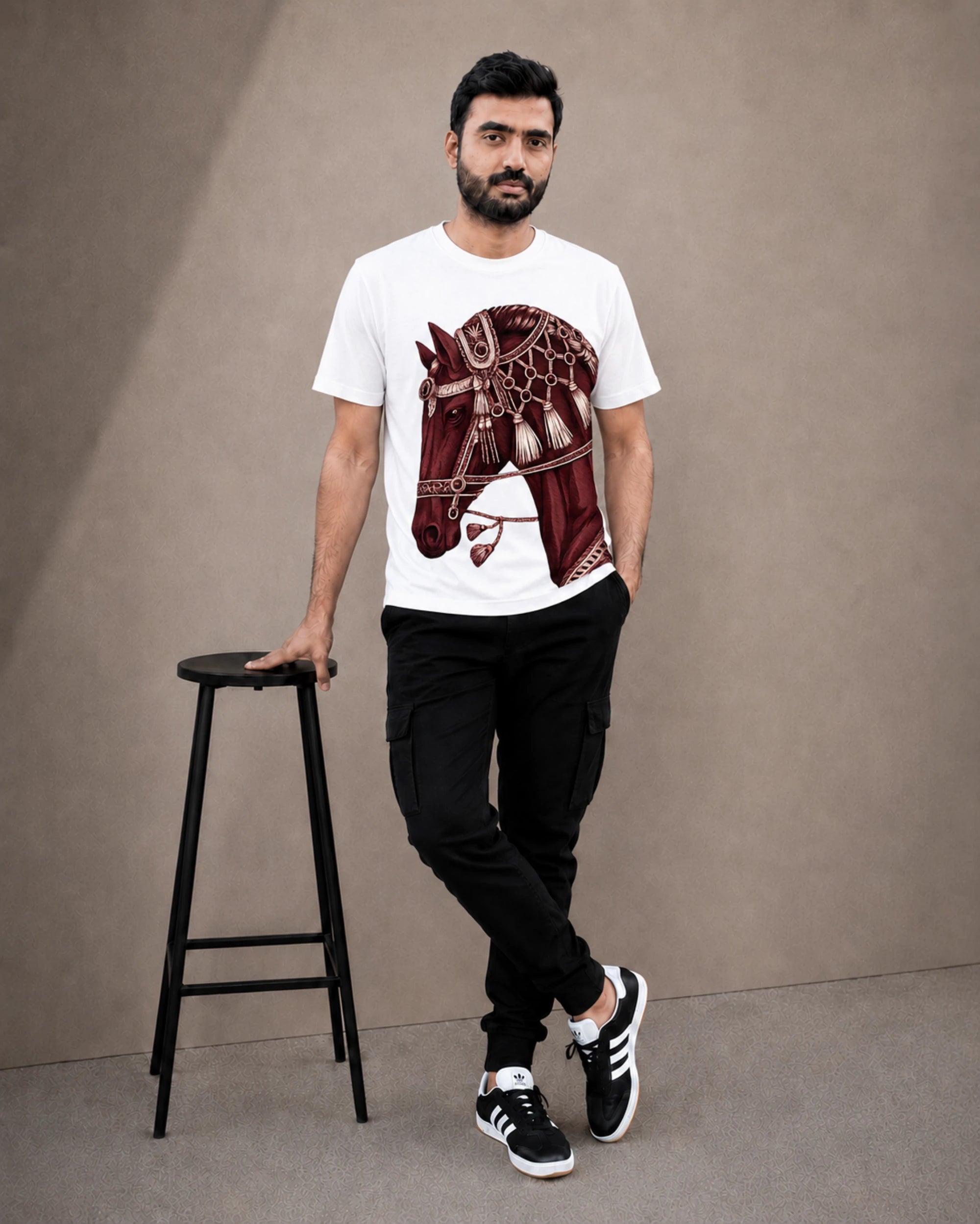 A man wearing a white crew-neck t-shirt with a detailed maroon horse graphic print, paired with black cargo pants and black-and-white sneakers, standing next to a black stool against a neutral studio backdrop.