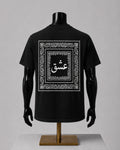 A black short-sleeve T-shirt displayed on a black mannequin torso with arms and a stand, shown from the back. The shirt features a large white ornamental border graphic with Arabic calligraphy in the center.