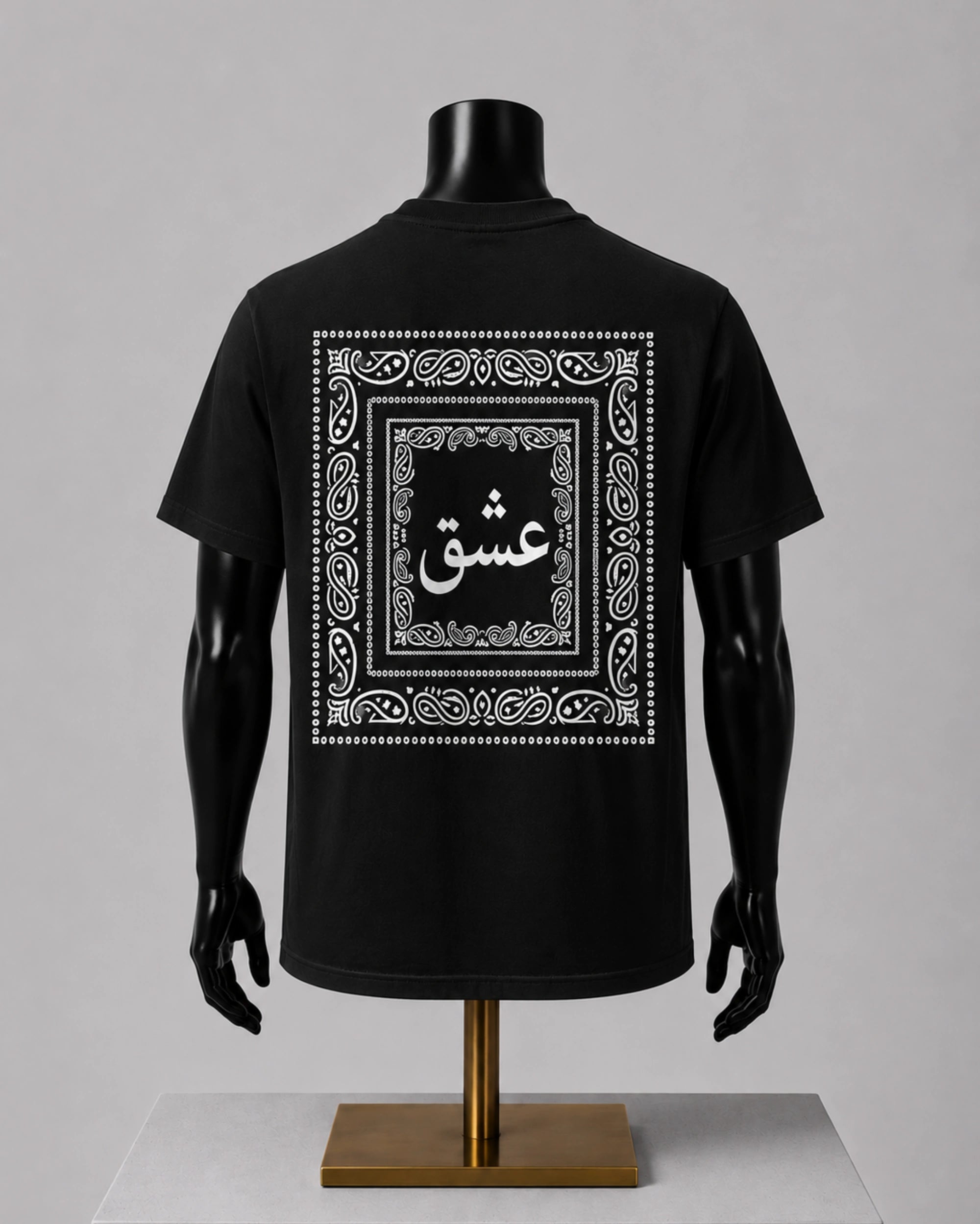 A black short-sleeve T-shirt displayed on a black mannequin torso with arms and a stand, shown from the back. The shirt features a large white ornamental border graphic with Arabic calligraphy in the center.