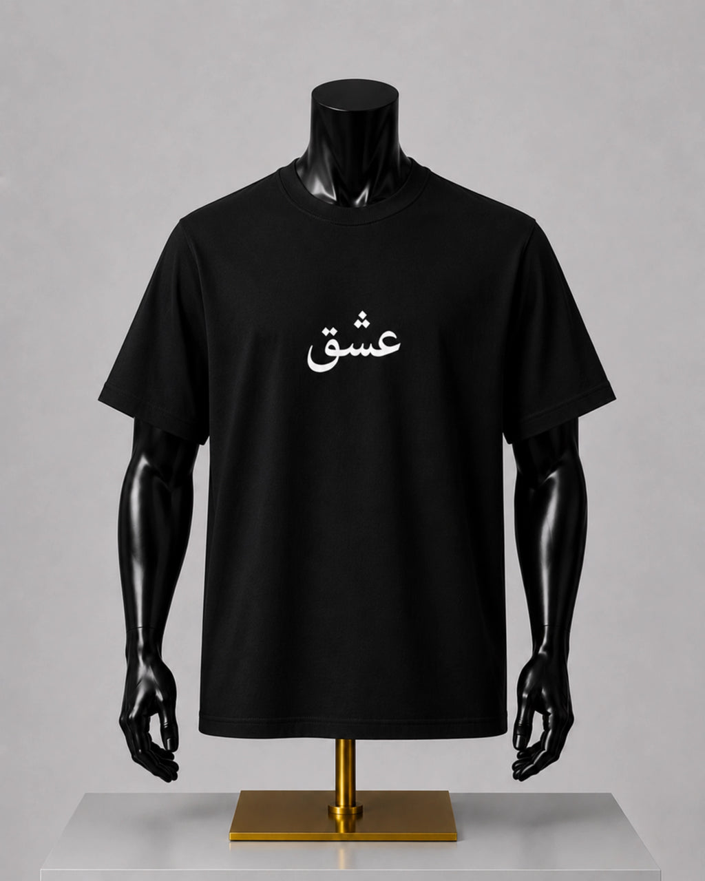 A black short-sleeve T-shirt displayed on a black mannequin torso with arms, mounted on a gold stand against a light gray studio background. The shirt has white Arabic script printed across the chest.