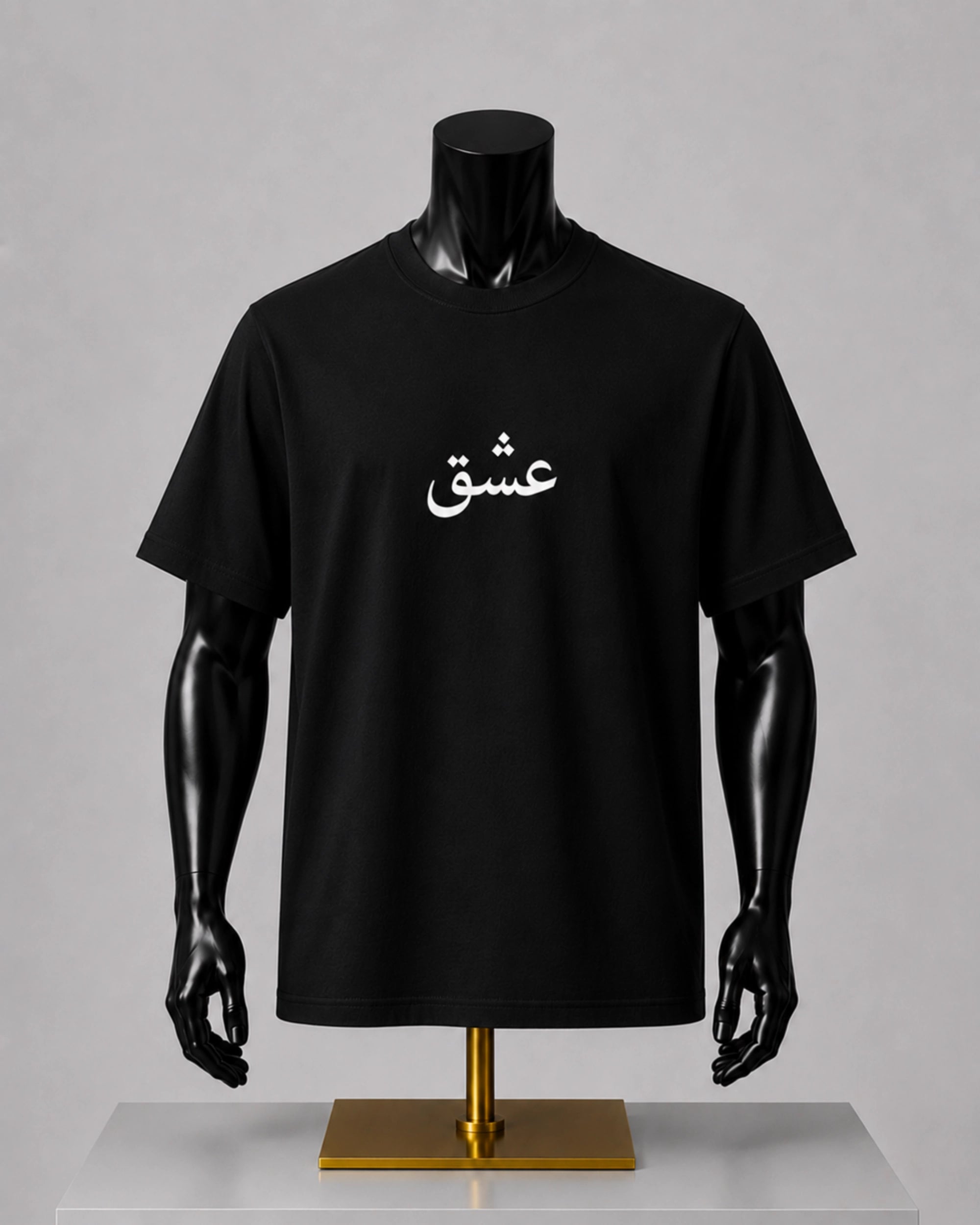 A black short-sleeve T-shirt displayed on a black mannequin torso with arms, mounted on a gold stand against a light gray studio background. The shirt has white Arabic script printed across the chest.
