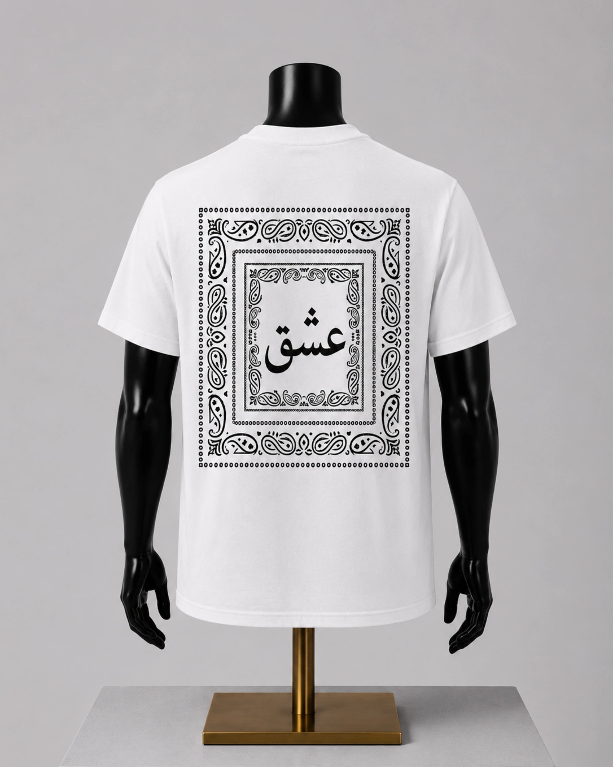 White short-sleeve T-shirt displayed on a black mannequin torso with a gold stand, shown from the back. The shirt features a large black ornamental border and Arabic calligraphy graphic centered on the back.