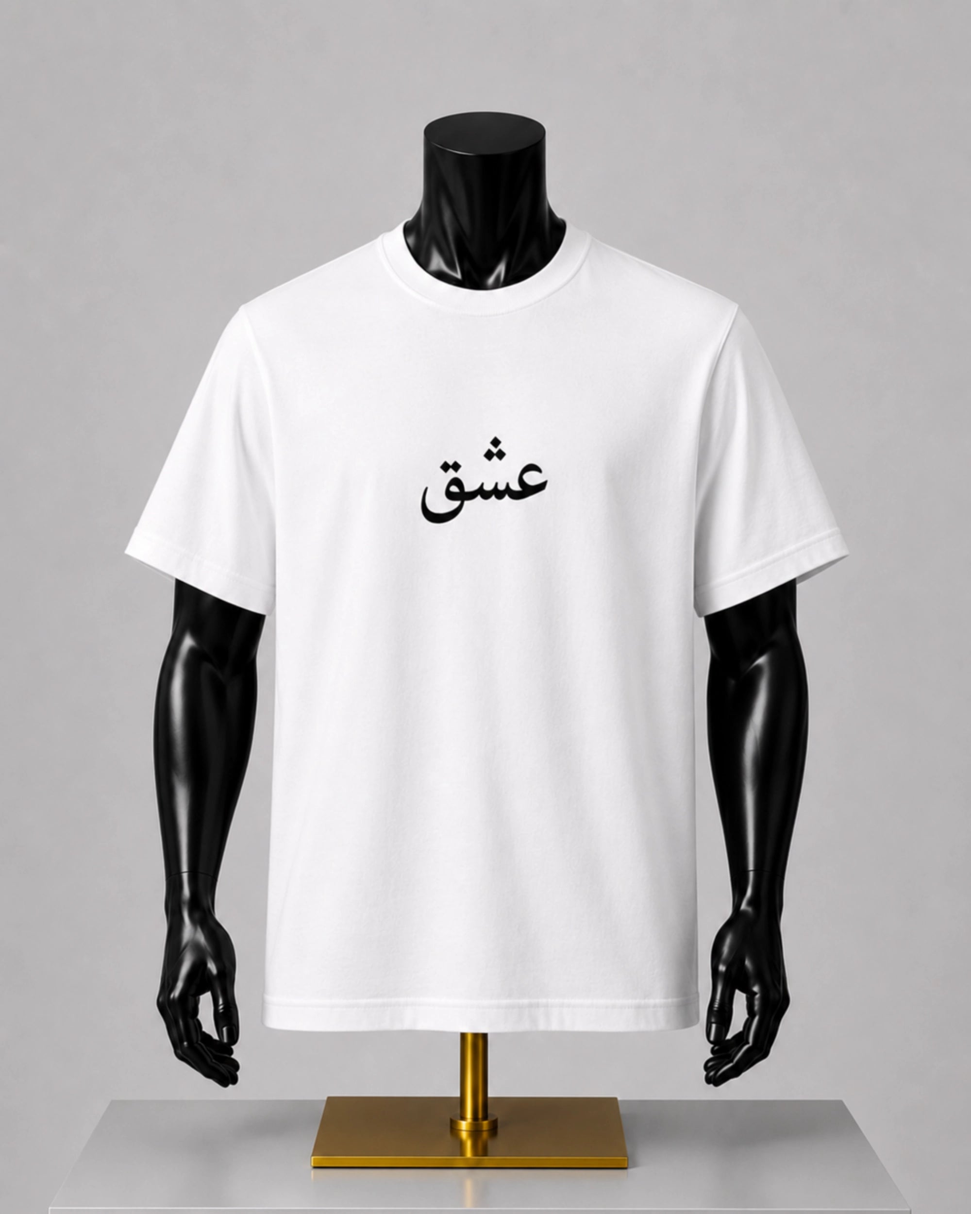A white short-sleeve crewneck t-shirt with black Arabic calligraphy printed on the chest, displayed on a black mannequin torso with a gold stand against a gray background.