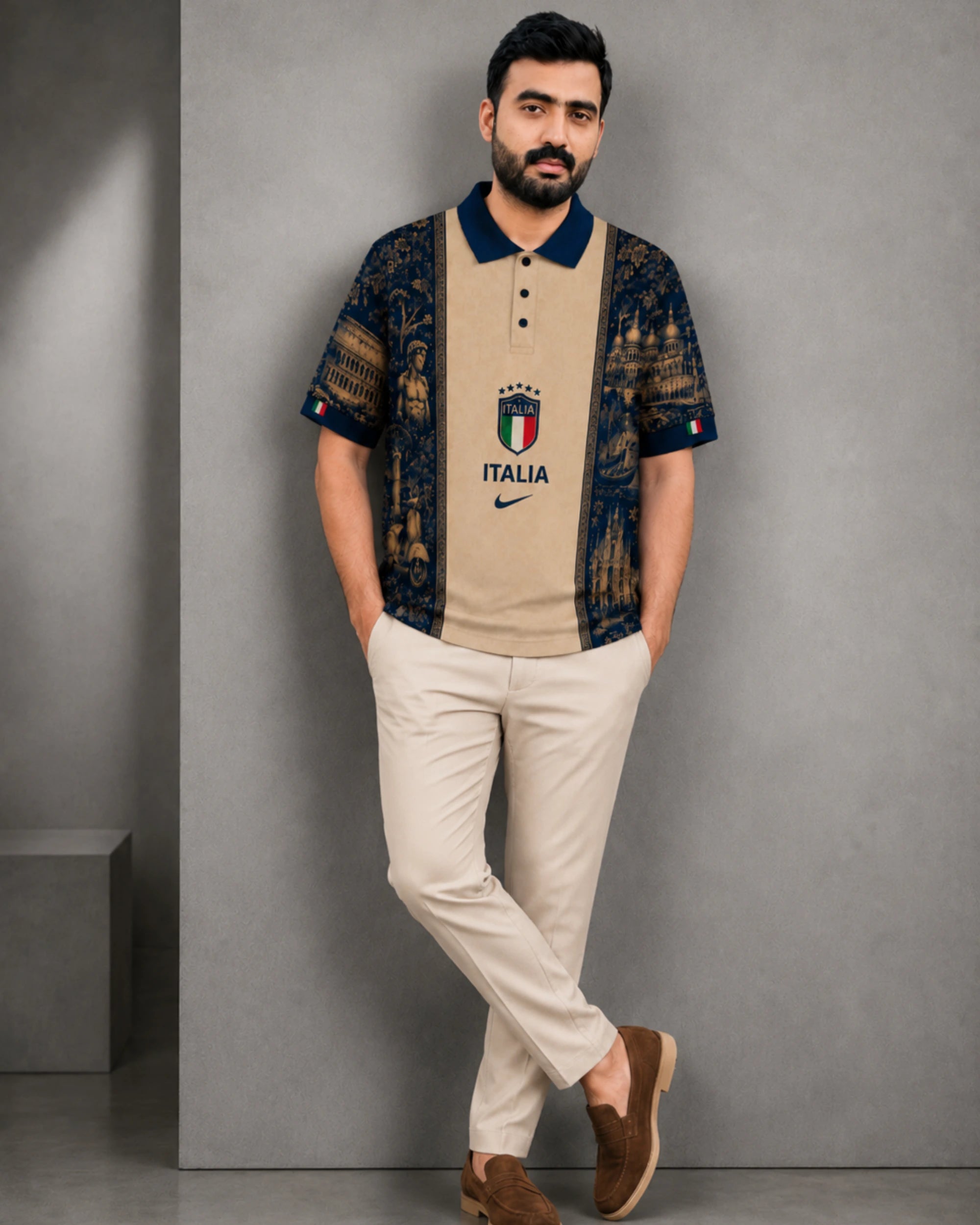 A man modeling a short-sleeve polo shirt with a beige center panel and dark navy blue patterned sleeves and collar, featuring an Italy crest and Nike logo. He is wearing beige trousers and brown loafers, standing against a gray concrete wall.