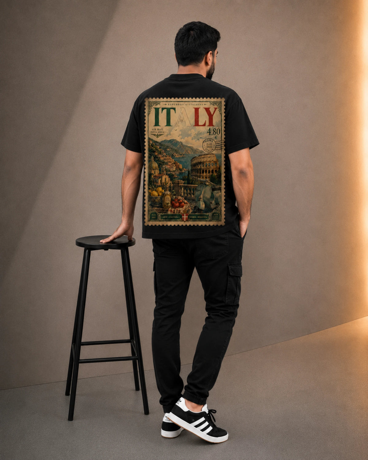 A man viewed from behind wearing a black t-shirt with a large vintage-style 'ITALY' travel poster graphic on the back, paired with black cargo pants and black-and-white sneakers, standing next to a black stool in a studio setting.